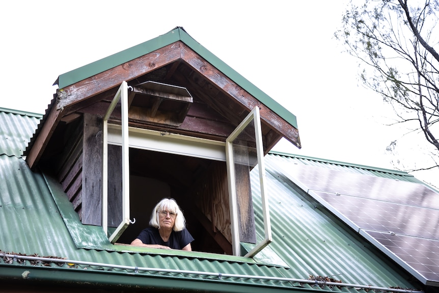 An older woman looks out from a window protruding from the roof of her two-storey house.