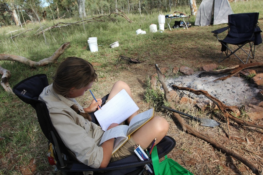 Woman sits in camping chair next to a campfire area, writing in a notebook.
