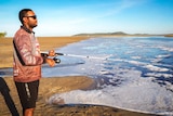 A man stands on a beach with sunnies on, looking out at the water holding a fishing rod. Clear blue skies.