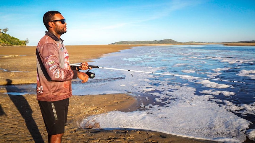 A man stands on a beach with sunnies on, looking out at the water holding a fishing rod. Clear blue skies.