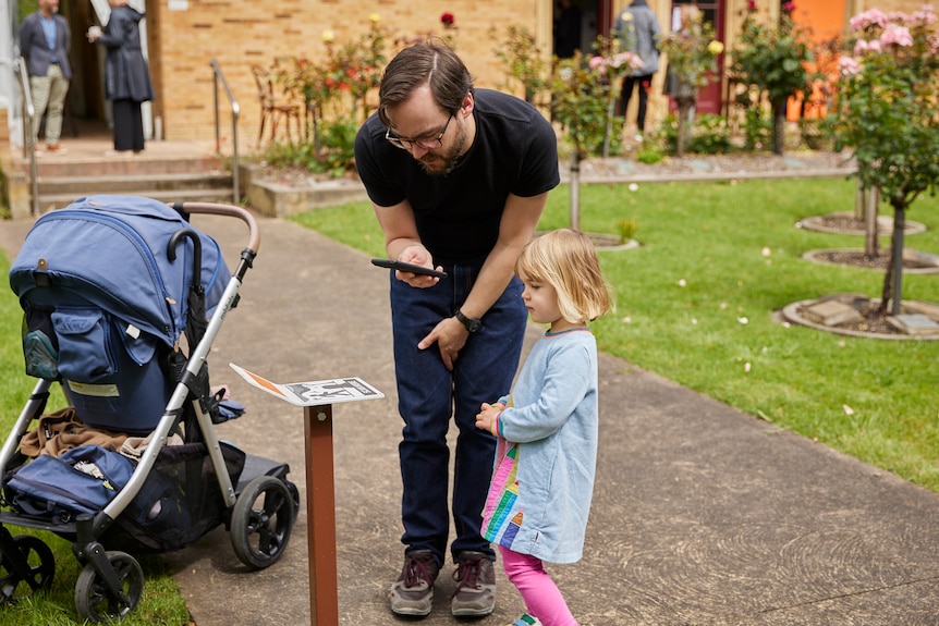Fraser Allison, in black shirt and pants, with a young child look at a smart phone held near a sign-post. Grass surrounds them.
