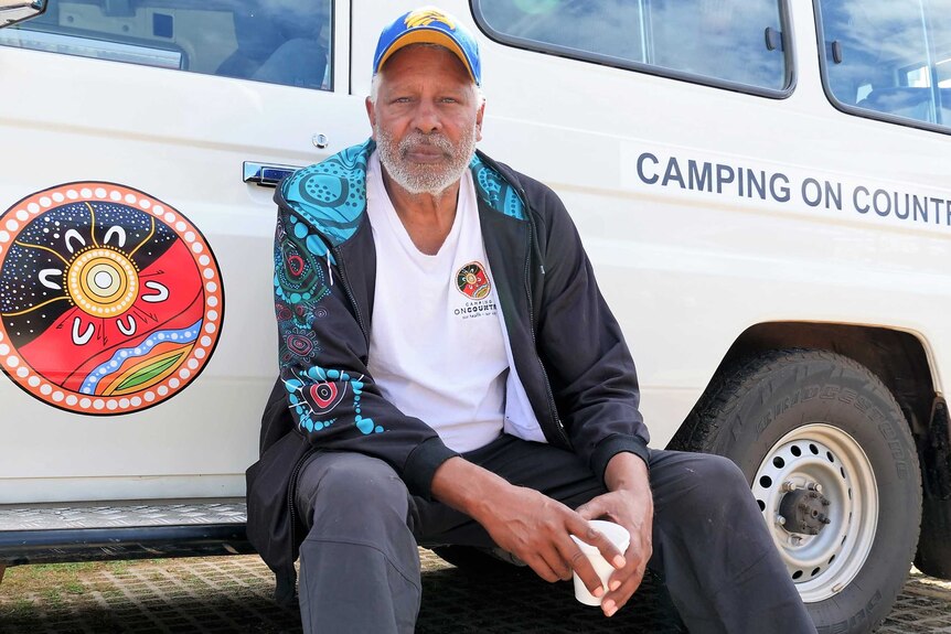 Aboriginal man in hat (Ernie Dingo) sitting on the step of a four-wheel-drive marked 'Camping On Country'.