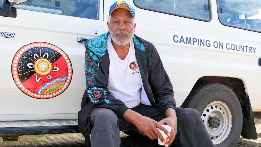 Aboriginal man in hat (Ernie Dingo) sitting on the step of a four-wheel-drive marked 'Camping On Country'.