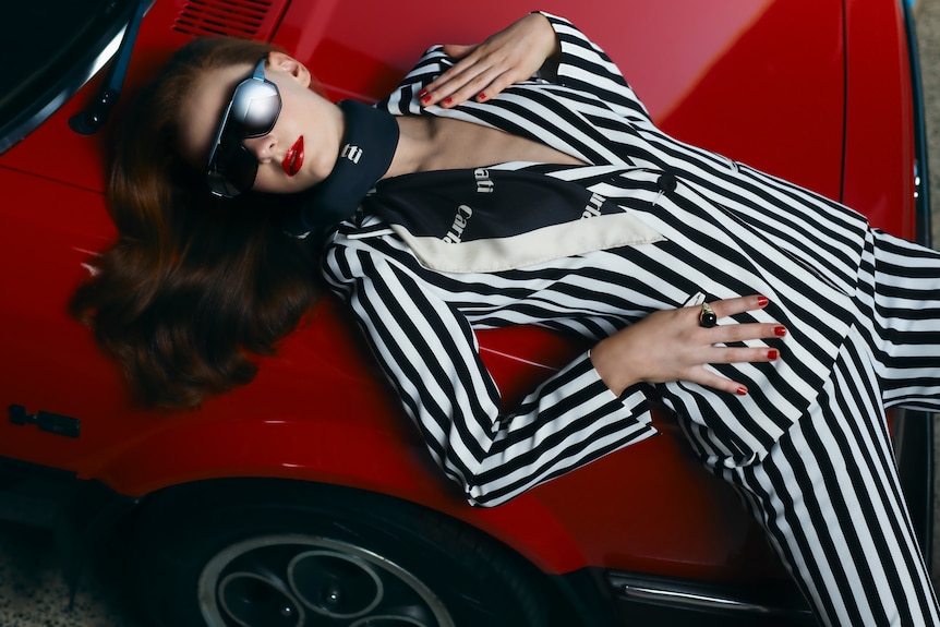 A female model with auburn hair wears dark glasses and a black and white striped suit while reclining on a red Ford.