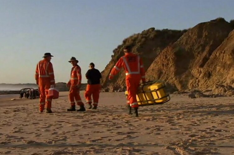 People in orange emergency services overalls walk on the beach towards a crowd of people.
