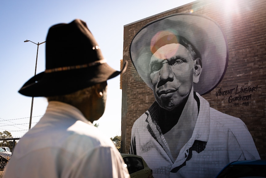 An Aboriginal man wears a wide-brimmed hat and looks up to a mural of Vincent Lingiari on a sunlit brick wall