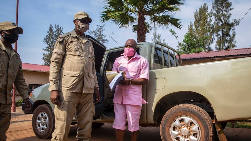 Paul Rusesabagina wears a pink prison suit as he stands near a truck with armed guards.