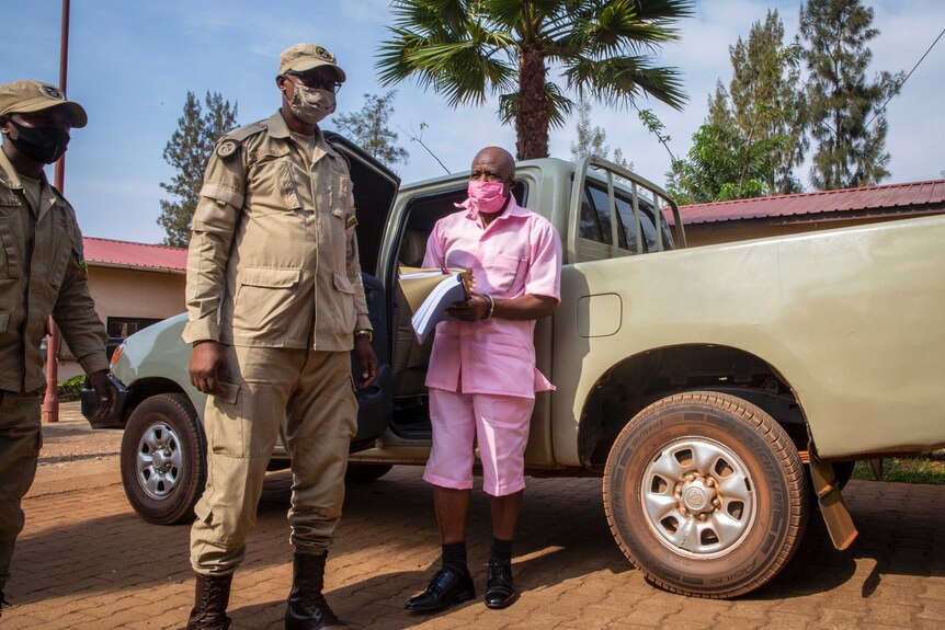 Paul Rusesabagina wears a pink prison suit as he stands near a truck with armed guards.