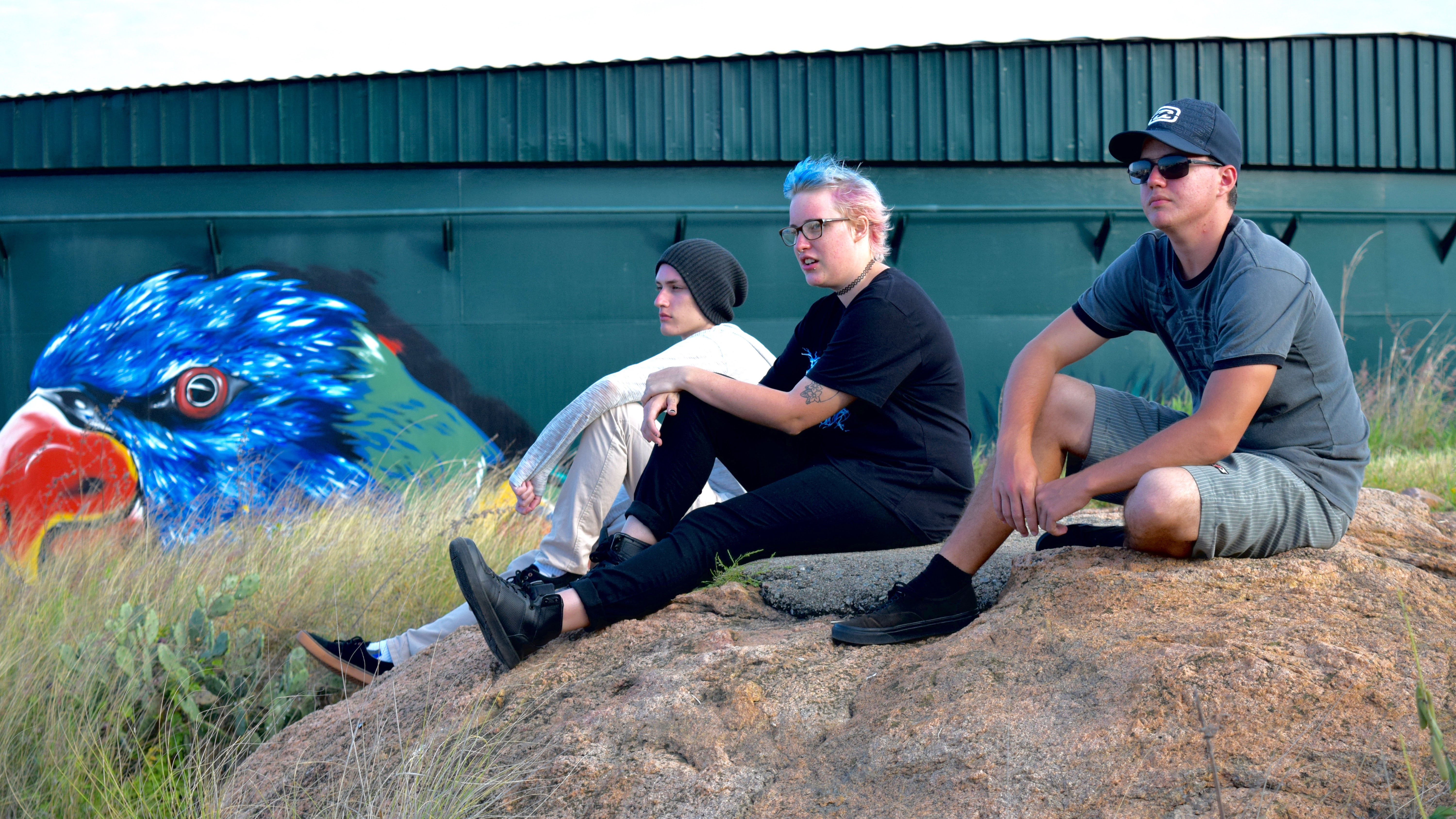 Three young people sit on a rock in front of a wall painted with graffiti.