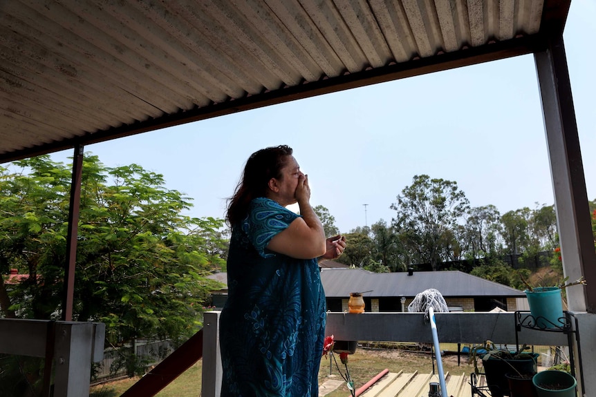 Woman in blue floral dress stands on shaded verandah with tin roof above and trees and houses in background