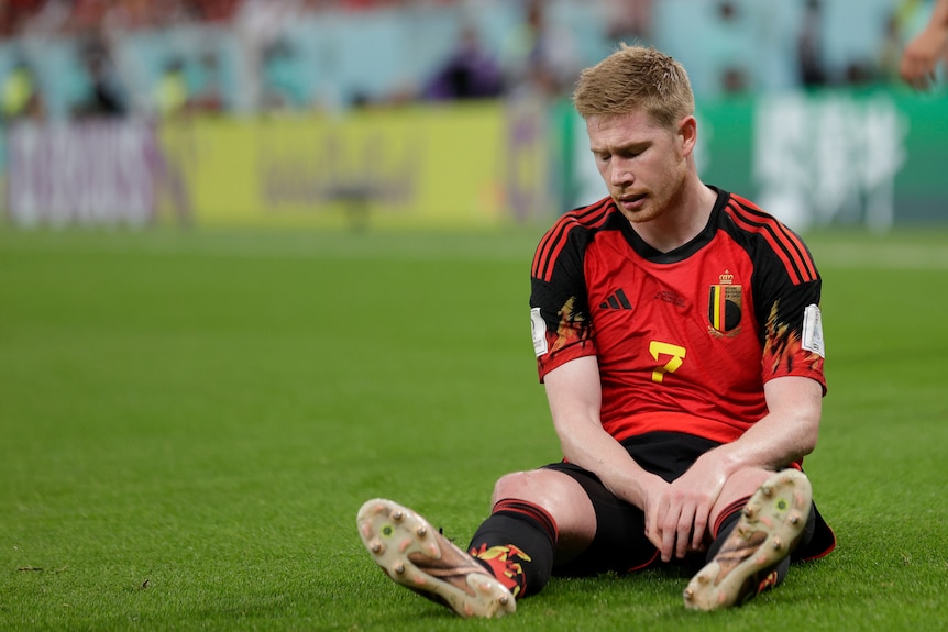 Kevin de Bruyne sits and looks down with his legs stretched out in front of him 