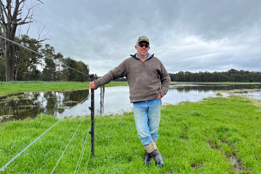A man stands in a paddock with a flooded pool behind him wearing a cap and a jumper. 