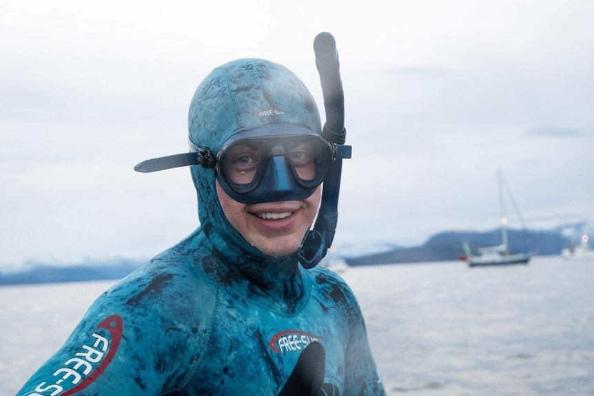 A man in a blue camoflauge wetsuit and snorkle gives the thumbs up with ocean in the background.