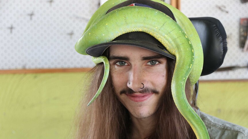 A young man with a snake wrapped around his baseball cap looks at the camera