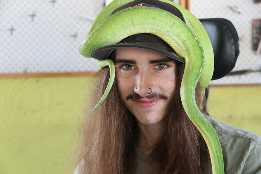 A young man with a snake wrapped around his baseball cap looks at the camera
