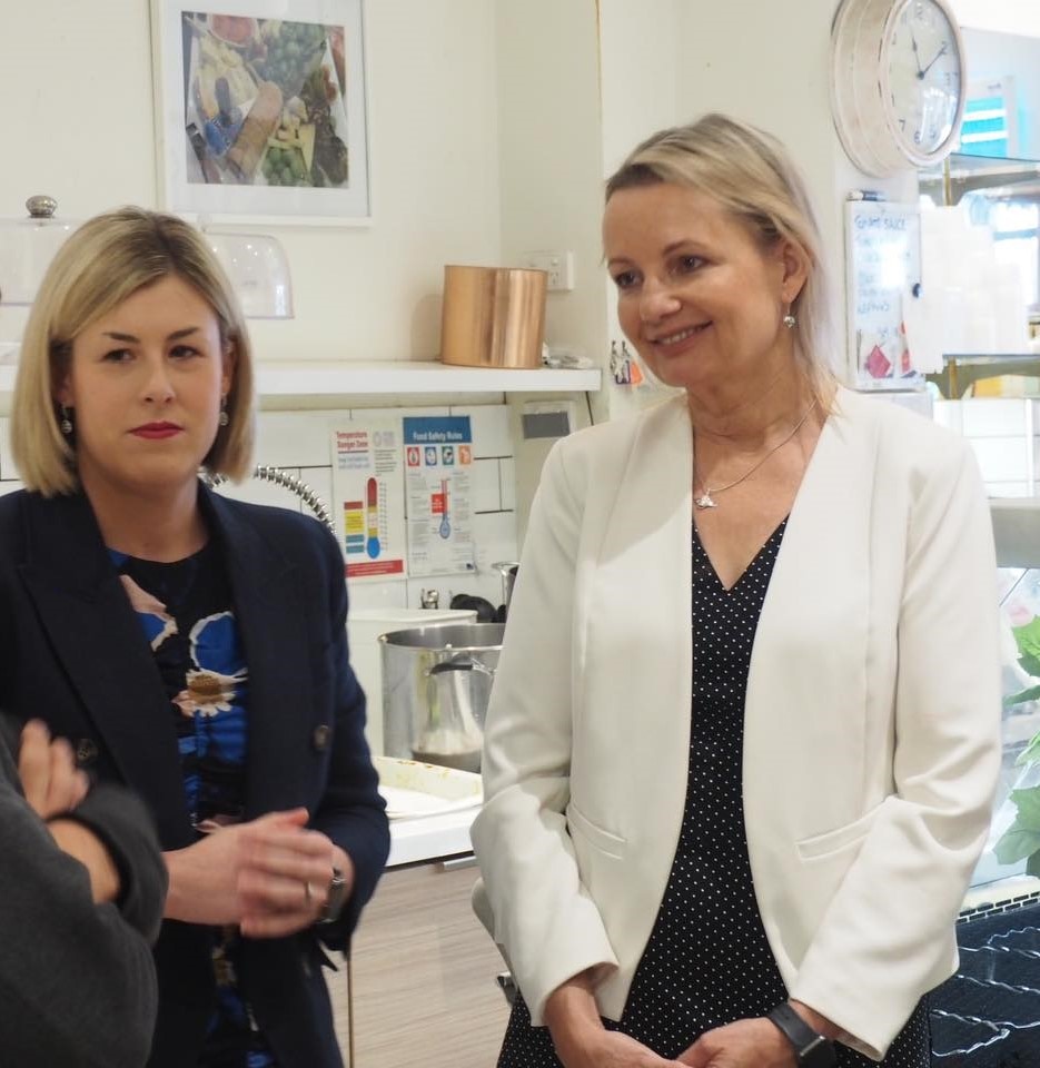 Two women with blonde hair stand in a cafe looking off camera.