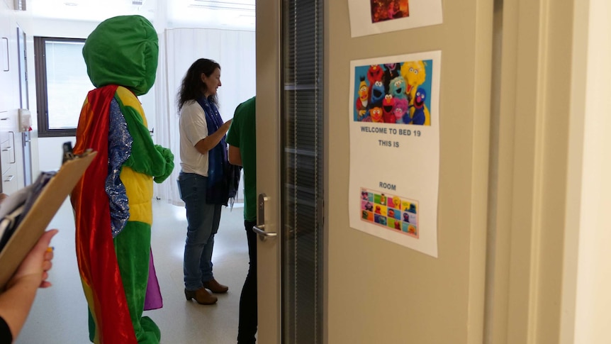 A person dressed as a giant turtle stands next to a woman, seen through an open door with the sign 'welcome to bed 19'.