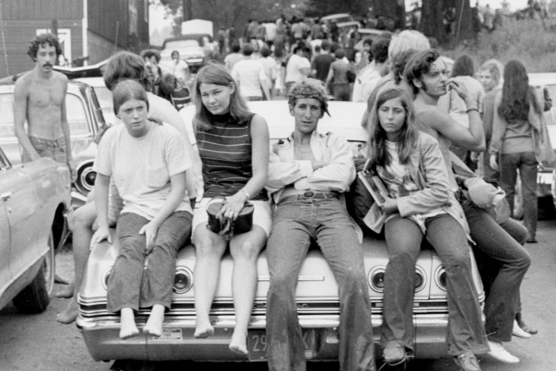 Young people sit on a car near, looking at the camera, surrounded by others.