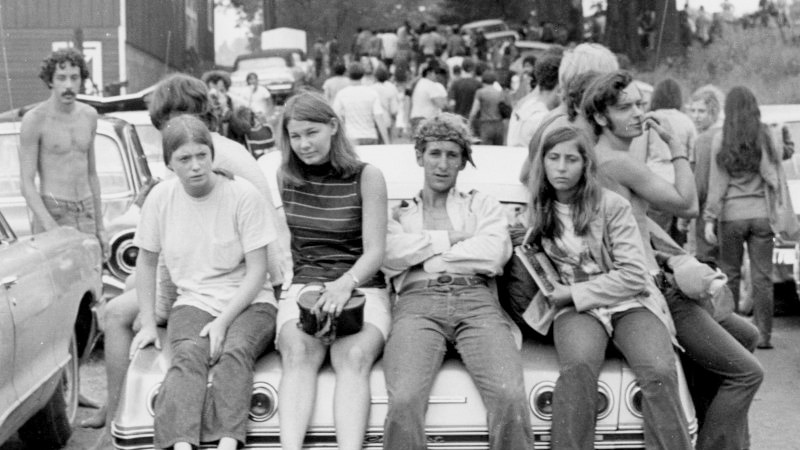 Young people sit on a car near, looking at the camera, surrounded by others