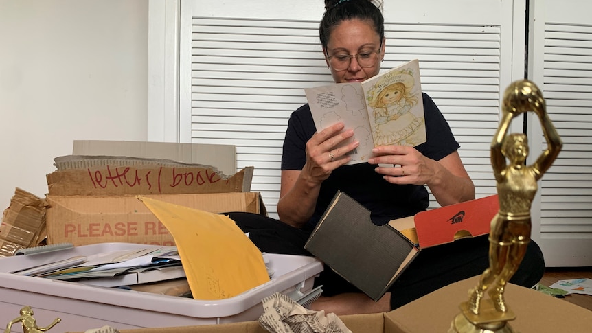 A woman surrounded by boxes, reading an old sixth birthday card. Sporting trophies and envelopes are among the open boxes.