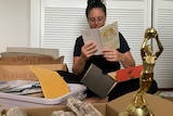 A woman surrounded by boxes, reading an old sixth birthday card. Sporting trophies and envelopes are among the open boxes.
