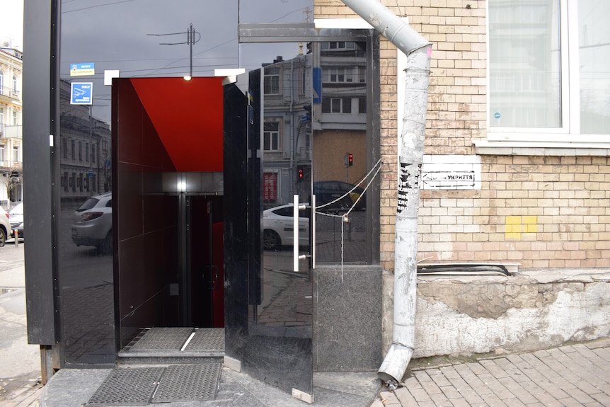 A black glass door leading to a set of stairs opens onto a Kyiv street, with dirty brick walls