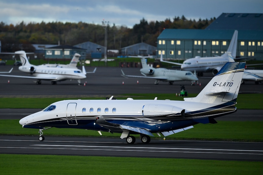 A number of private jets and small planes sit on tarmac at an airport. Large and small buildings in the background.