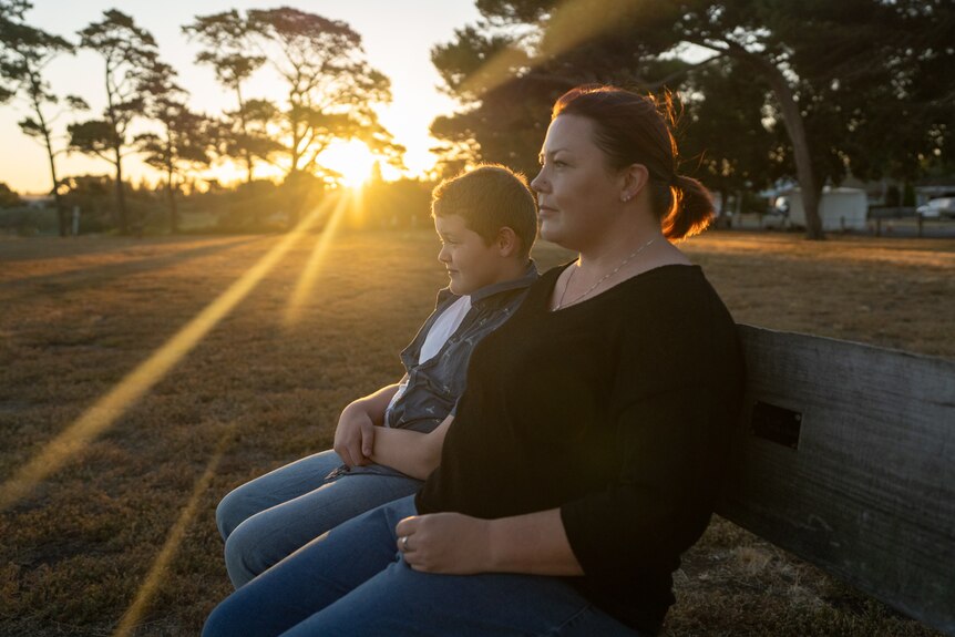 A boy and his mum sit on a park bench looking ahead. To their right the sun is setting behind a row of trees.