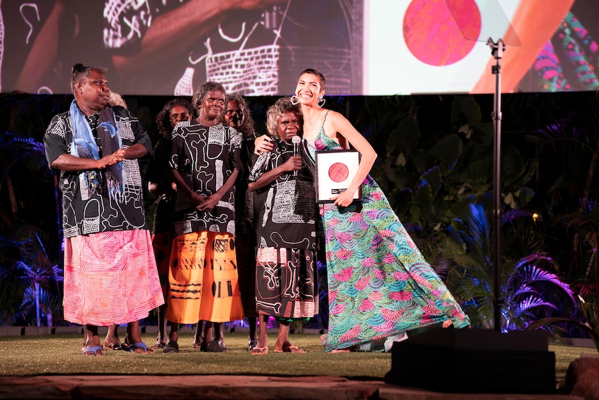 A group of women stood on stage.  One hugs a woman with a microphone.