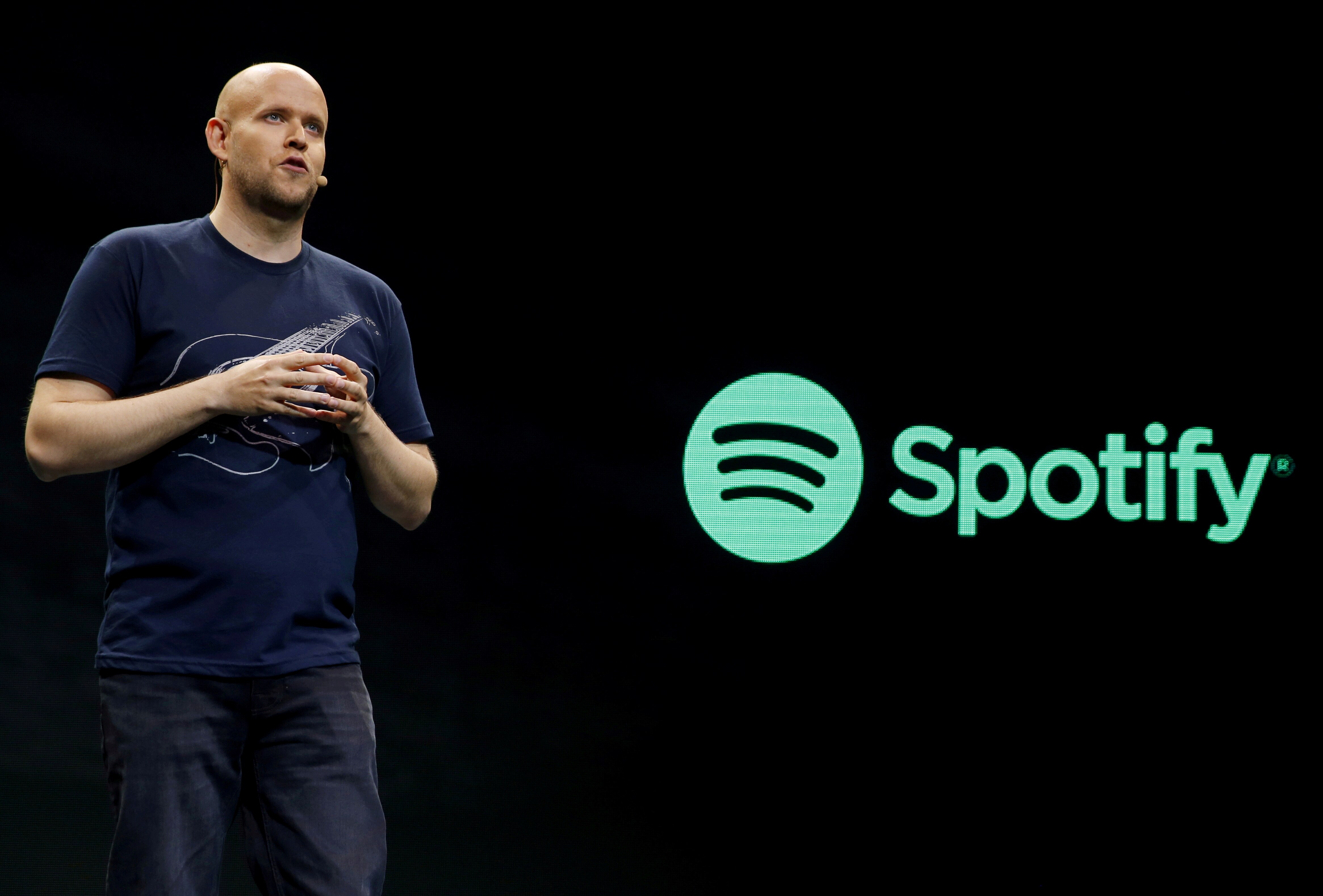 A man with a microphone piece is standing in front of a board which has the Spotify logo. 
