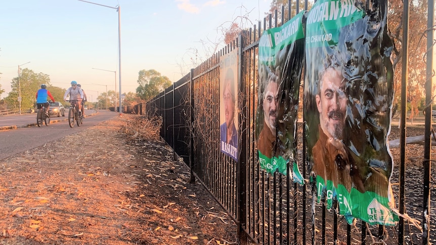 two singed plastic posters on a fence as cyclists pass on a nearby pathway.