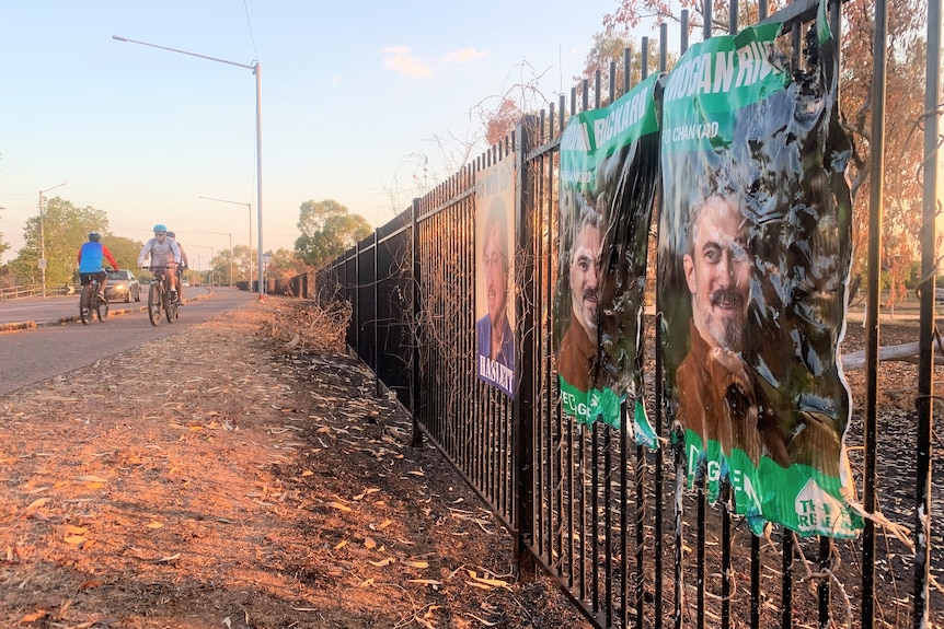 two singed plastic posters on a fence as cyclists pass on a nearby pathway.