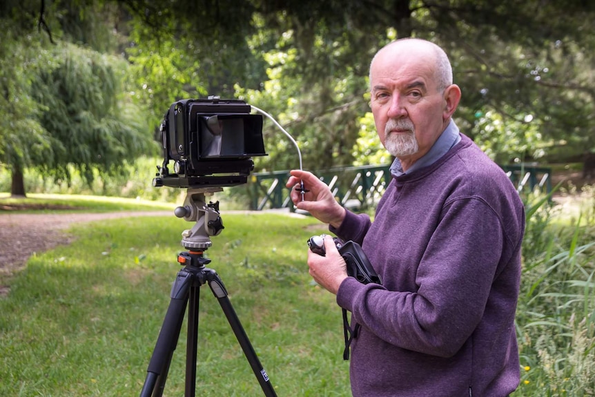 A man stands next to an analogue camera and holds the cord with the button used to take the photograph