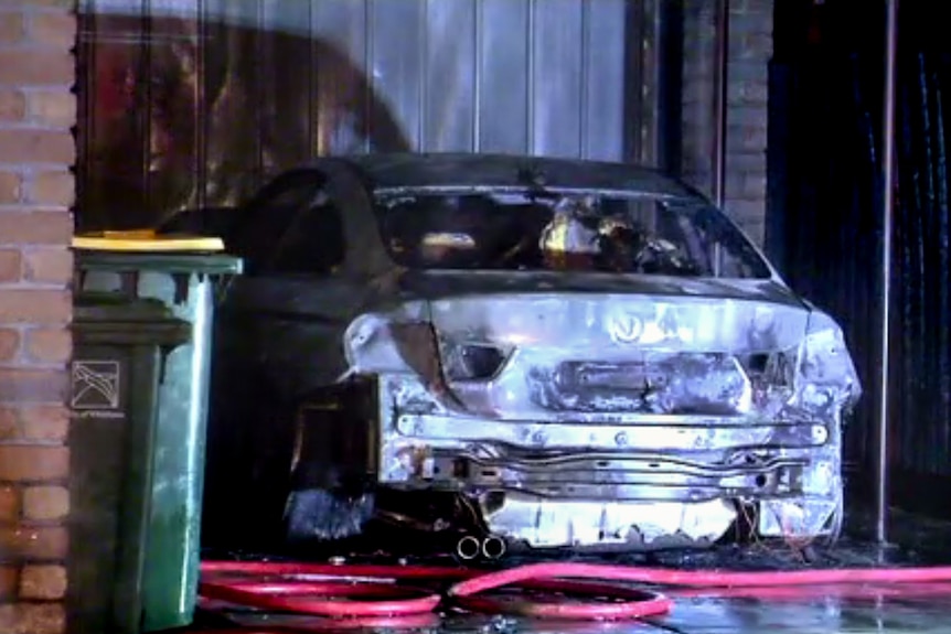 A BMW sits burnt out in a driveway, with  a brick wall and wheelie bin next to it.