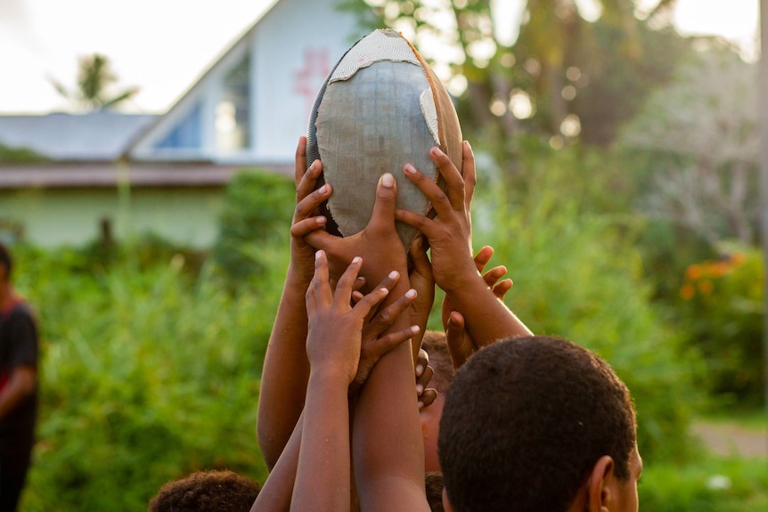 Several hands of young boys hold up a battered rugby ball