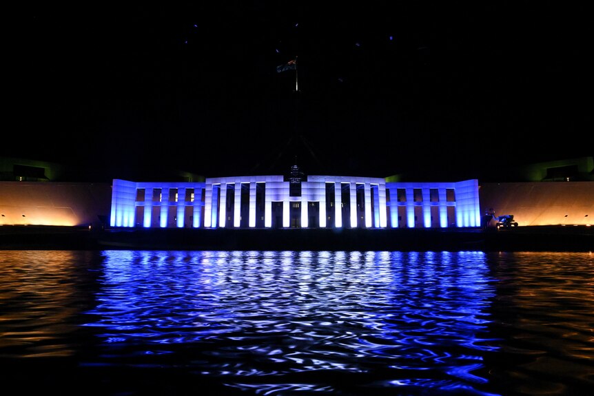 A wide view of the front of Parliament House across the water shows it illuminated in blue and white colours at night.