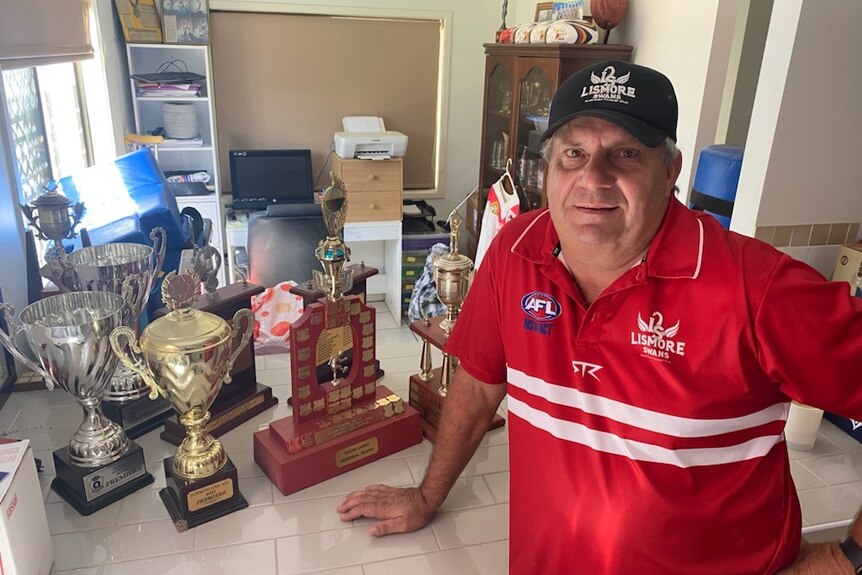 A man wearing a red football guernsey stands in a home kitchen. On the bench next to him is multiple football trophies.