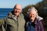 Don and Gail, dressed in warm jackets, smile as they stand in front of the sea on a sunny day.