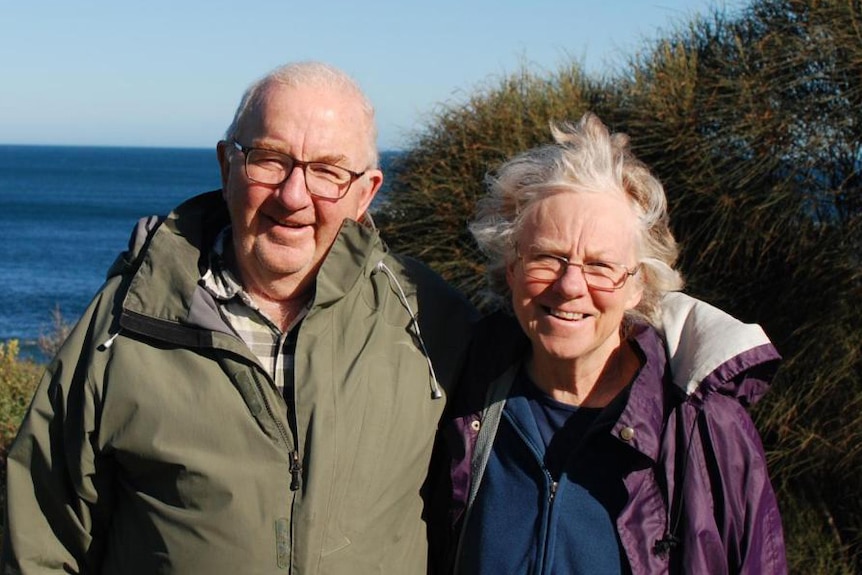 Don and Gail, dressed in warm jackets, smile as they stand in front of the sea on a sunny day.