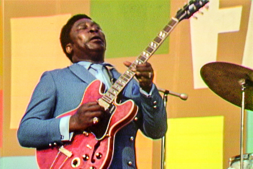 BB King in a sky blue suit with matching shirt and tie closes his eyes playing a red guitar against bright geometric wallpaper.