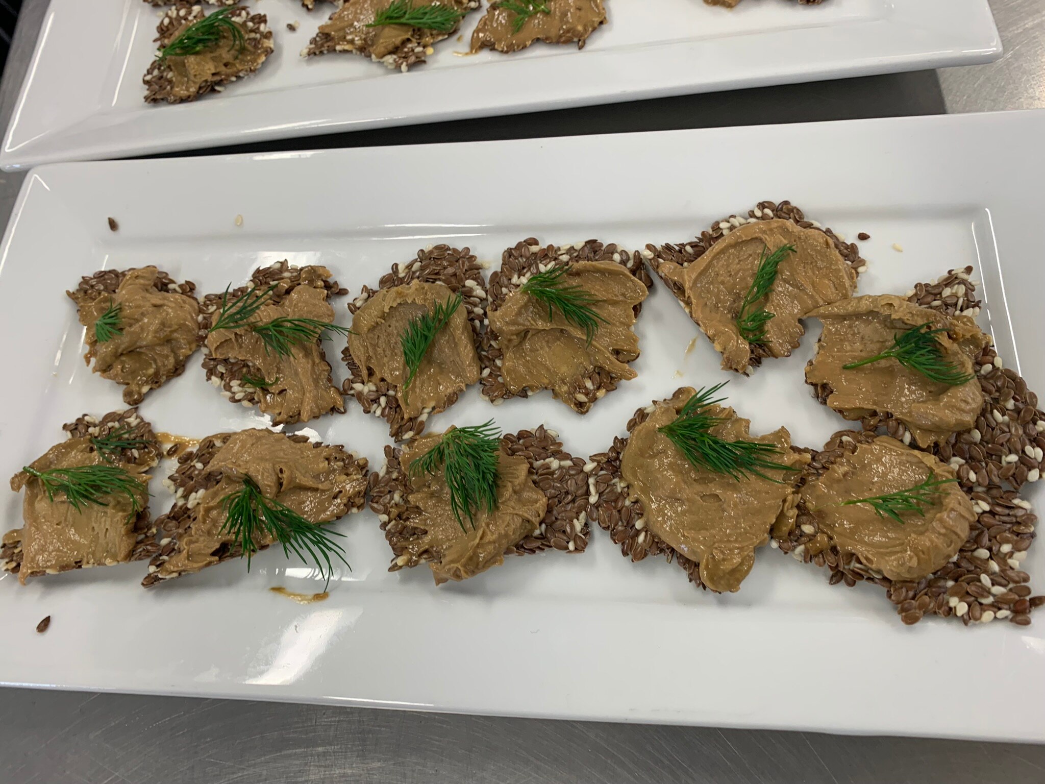 Photo of pate on handmade crackers. They are topped with sprigs of dill and are set out on a white, oblong china platter.