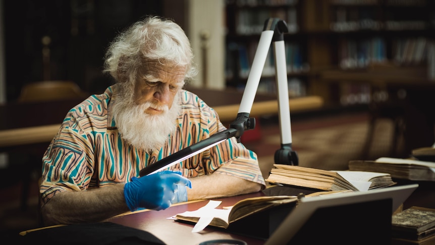 An older man with white hair and full beard wears gloves to read a book.