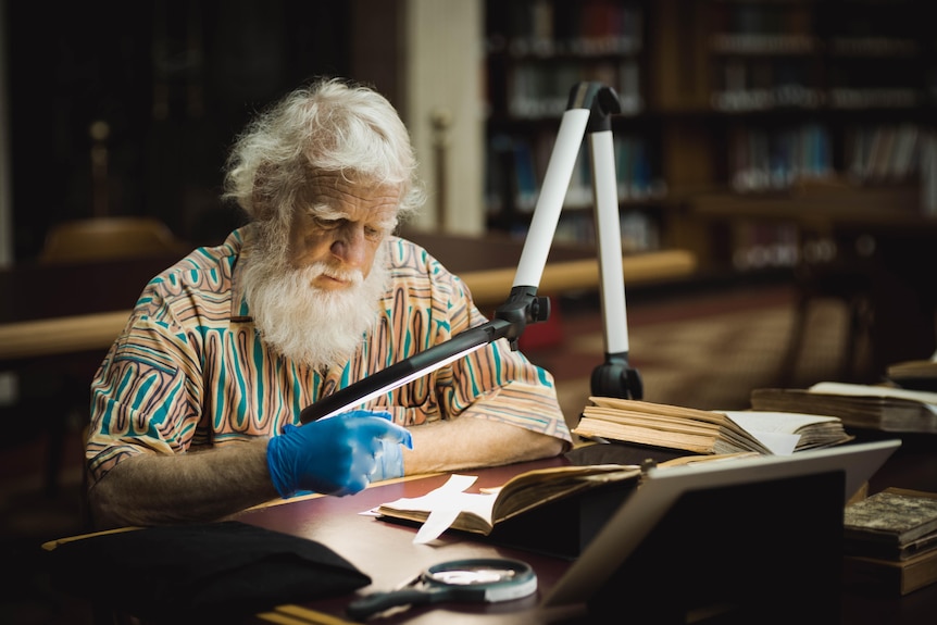 An older man with white hair and full beard wears gloves to read a book.