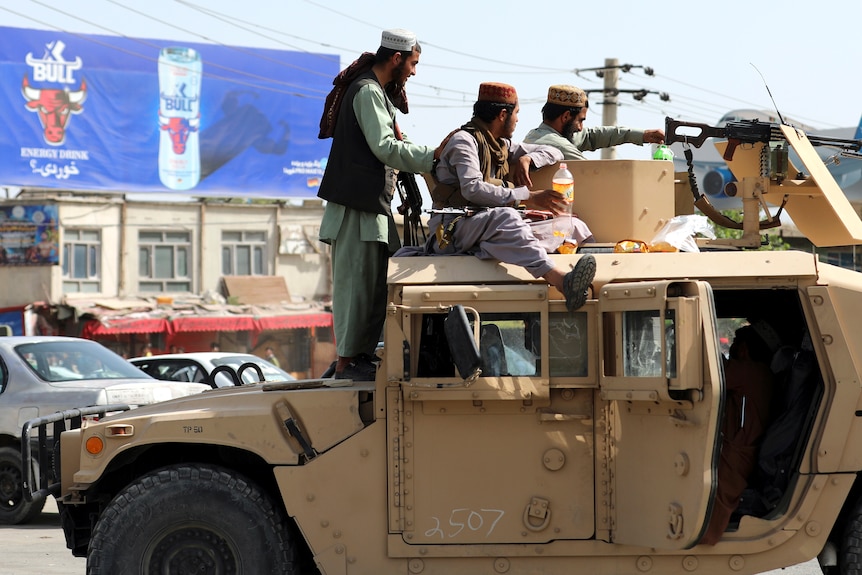 Three armed Middle Eastern men in traditional garb sit on top a yellow vehicle in city street.