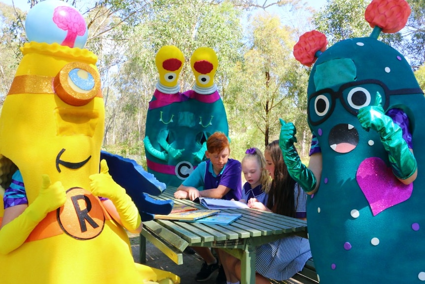 Three people in costumes surround three young school students, who are reading