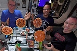 Five astronauts in t-shirts gather around a makeshift table in the space station with four pizzas floating in the air.