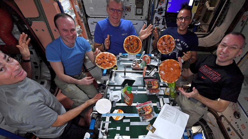 Five astronauts in t-shirts gather around a makeshift table in the space station with four pizzas floating in the air.
