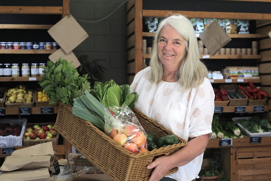  Woman with long hair in a white shirt holds a woven basket of apples and leaf greens. Behind her are grocery shelves