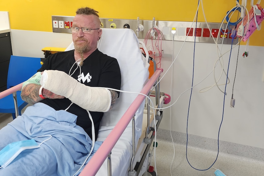 An image of Steve Whiteley in a hospital bed with a brace on his arm surrounded by hospital machinery and cords