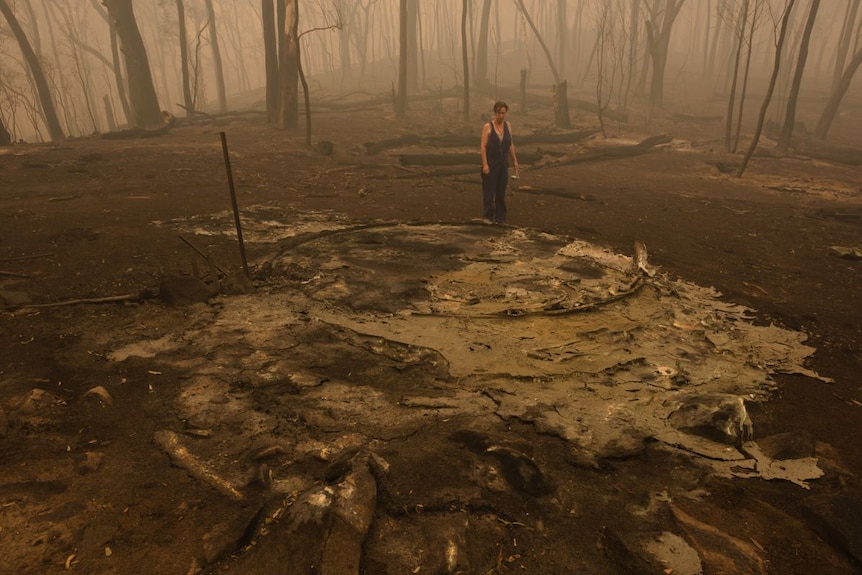 A woman stands by the plastic remnants of a water tank in a smoky forest.